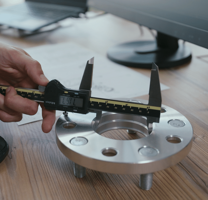 Digital caliper measuring a metal flange on a wooden desk near a computer.