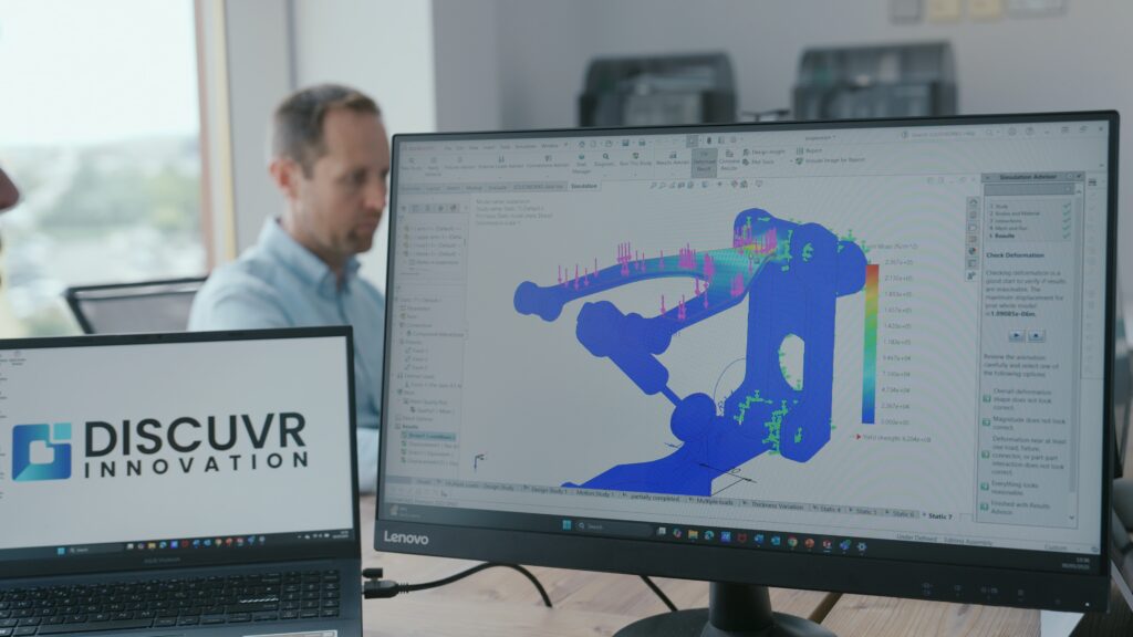 Two computer screens showing engineering software and DISCUVR Innovation logo, with a person working in the background.