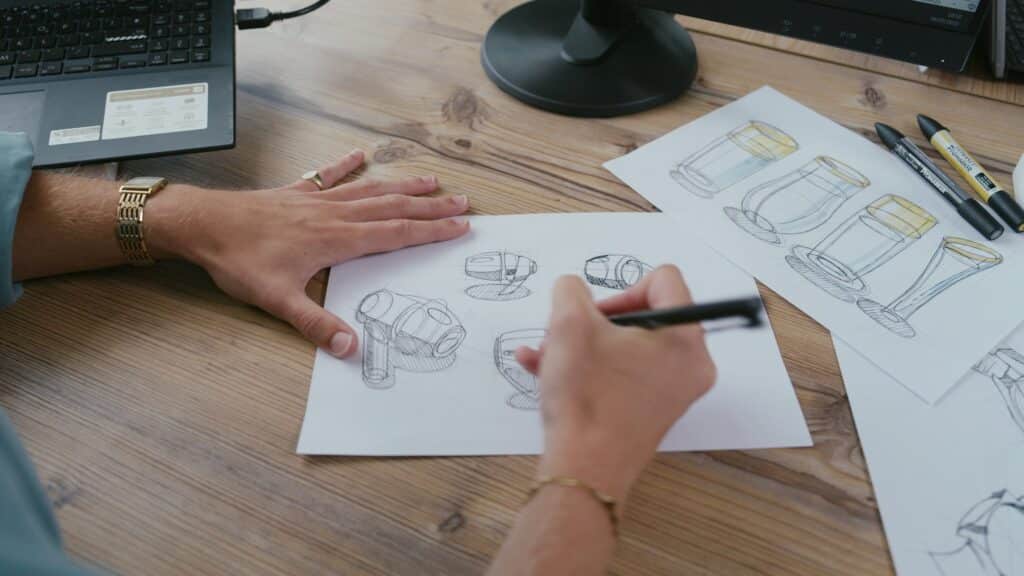 Person sketching industrial design concepts at a desk with drawing tools and a laptop.