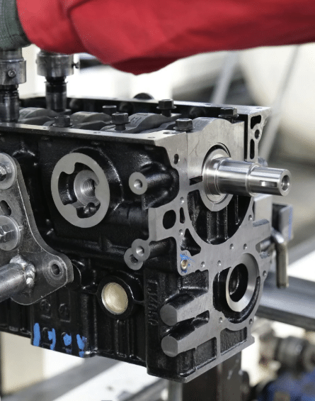 Engine block on an assembly line with mechanical components and a worker's arm in a red sleeve.