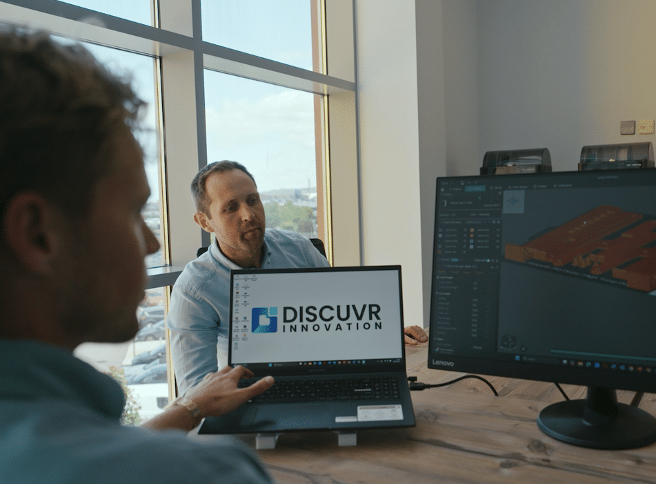 Two men discussing a project on a laptop displaying "DISCUVR Innovation" with a 3D model on a monitor.