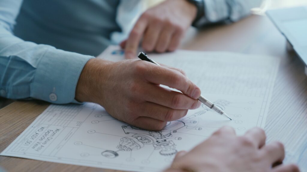 Hands reviewing technical drawing with a pen on a wooden table.
