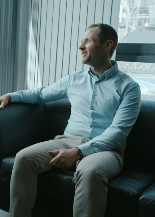 Person sitting on a black couch in a light blue shirt, looking out the window.